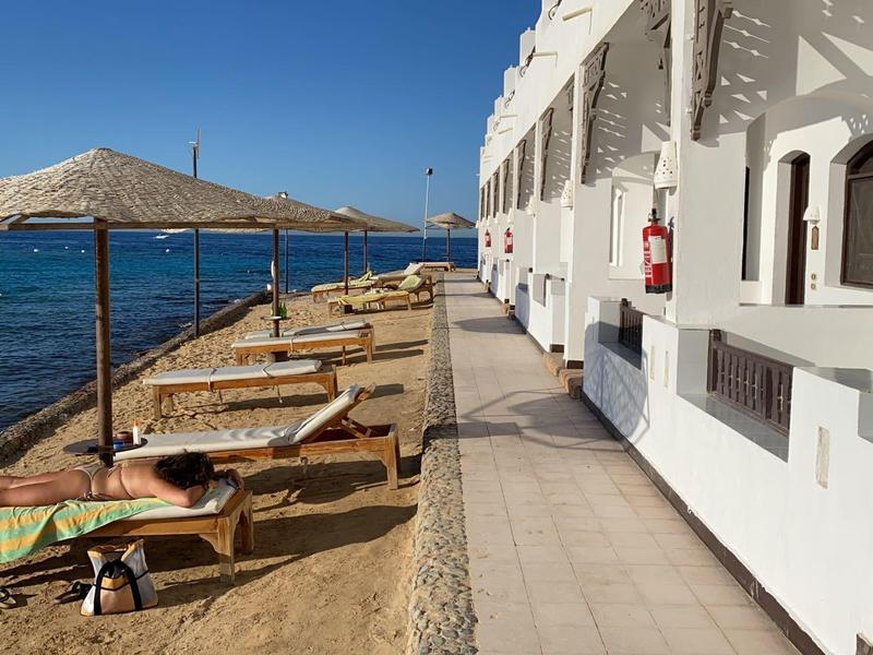Hotelterrasse mit Liegestühlen und Sonnenschirmen neben dem Meer unter blauem Himmel.