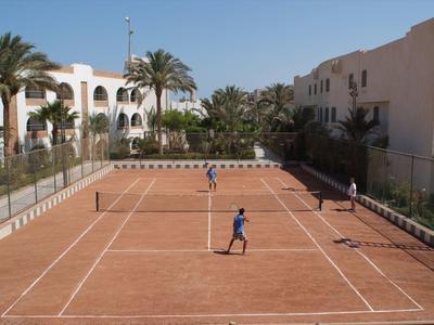 Ein roter Tennisplatz mit zwei Spielern, umgeben von hellen Gebäuden und Palmen unter blauem Himmel.