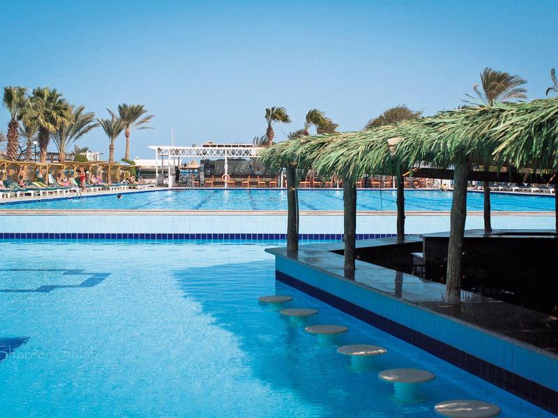 Großer Hotelpool mit Palmen und überdachter Bar im Freien unter blauem Himmel.