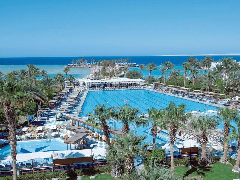 Großer Außenpool mit Liegestühlen und Palmen in einem Hotelresort am Meer.