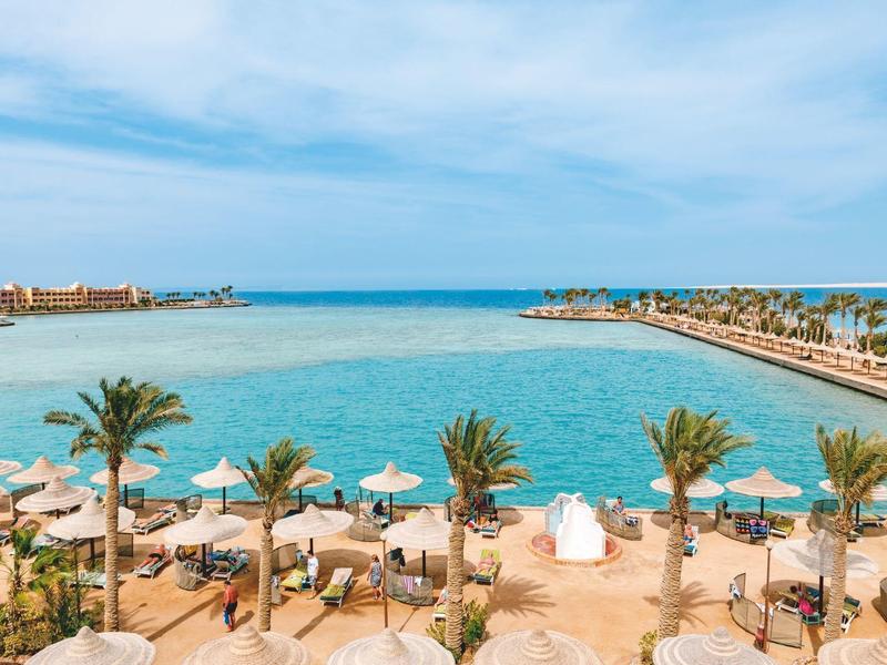 Strand mit Liegestühlen und Sonnenschirmen neben ruhigem Meer bei blauem Himmel.