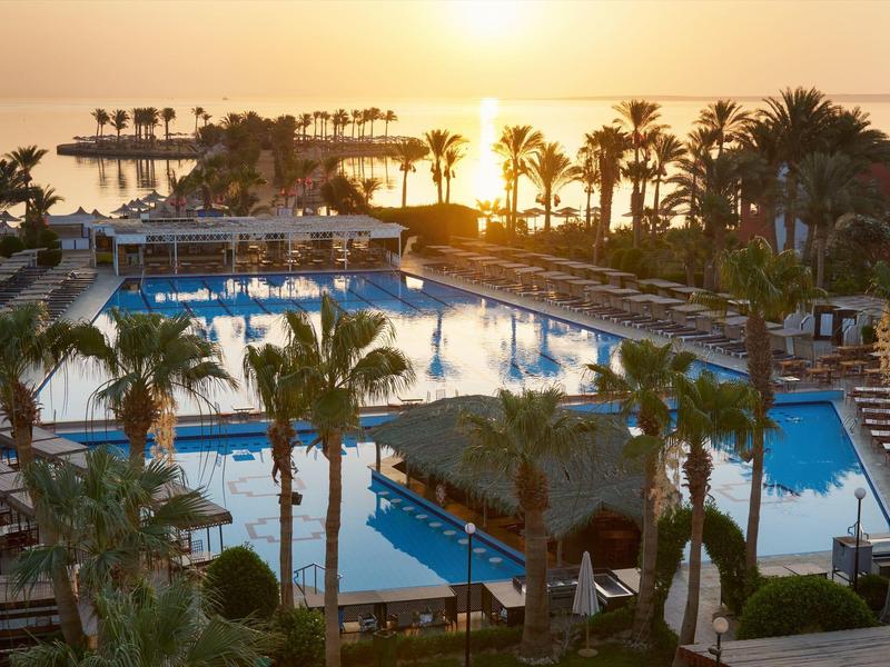 Ein großer Poolbereich mit Palmen und Sonnenuntergang am Meer im Hintergrund.