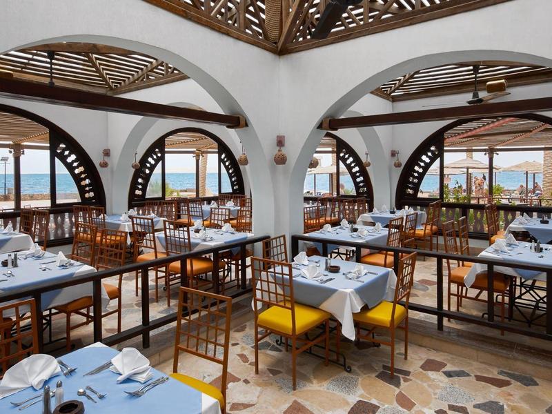 Helles Restaurant mit Tischen, Stühlen und Blick auf das Meer durch große Fenster.