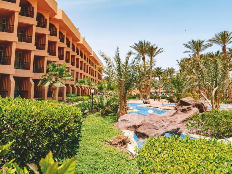 Hotelgebäude mit Balkonen neben einem Garten und Poolbereich mit Palmen.