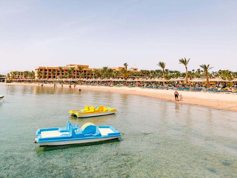 Leere Tretboote auf klarem Wasser vor einem Sandstrand mit Palmen und Gebäuden im Hintergrund.