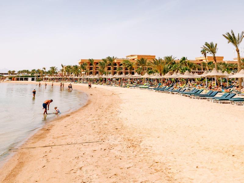 Breiter Sandstrand mit Liegen, Menschen und Palmen vor einem Hotel bei sonnigem Wetter.