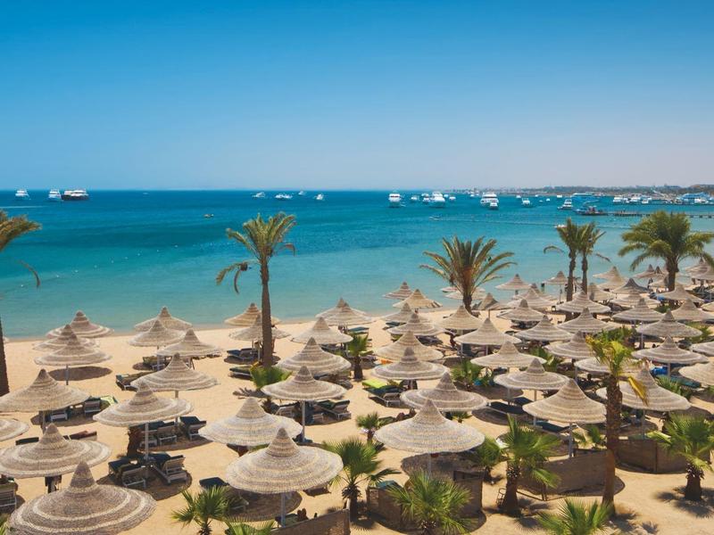 Sandstrand mit vielen Sonnenschirmen und Palmen, Blick auf ruhiges, blaues Meer mit Booten.