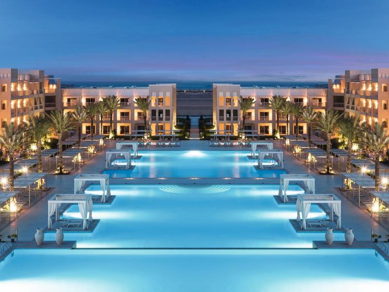 Großer, beleuchteter Pool umgeben von Hotelgebäuden und Pflanzen bei Abenddämmerung.