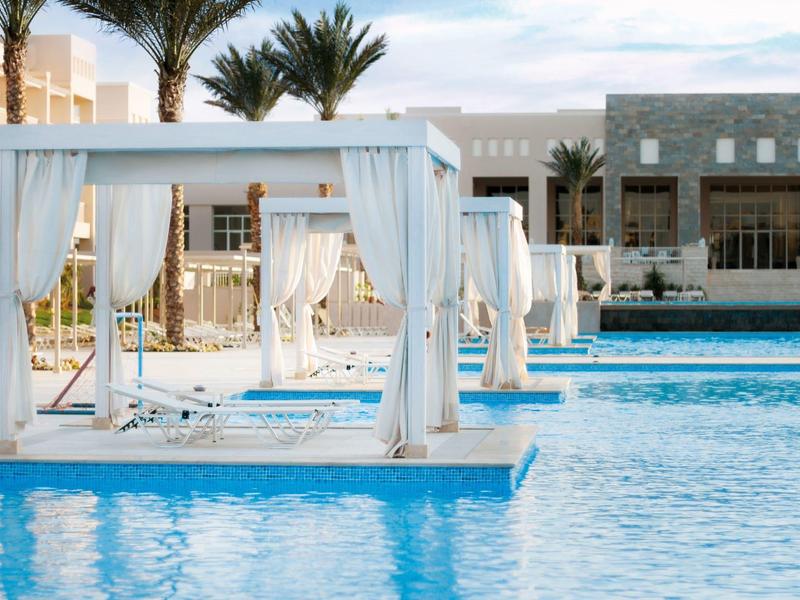 Weißes Poolbett mit Vorhängen, klarer blauer Pool und Palmen vor modernen Gebäuden.