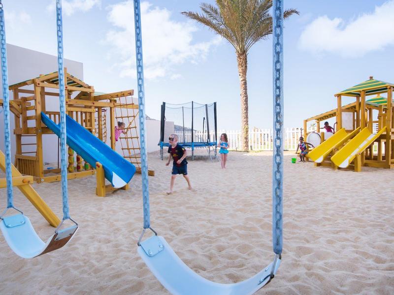 Spielplatz am Strand mit Schaukeln, Rutschen, Sandboden und spielenden Kindern bei klarem Himmel.