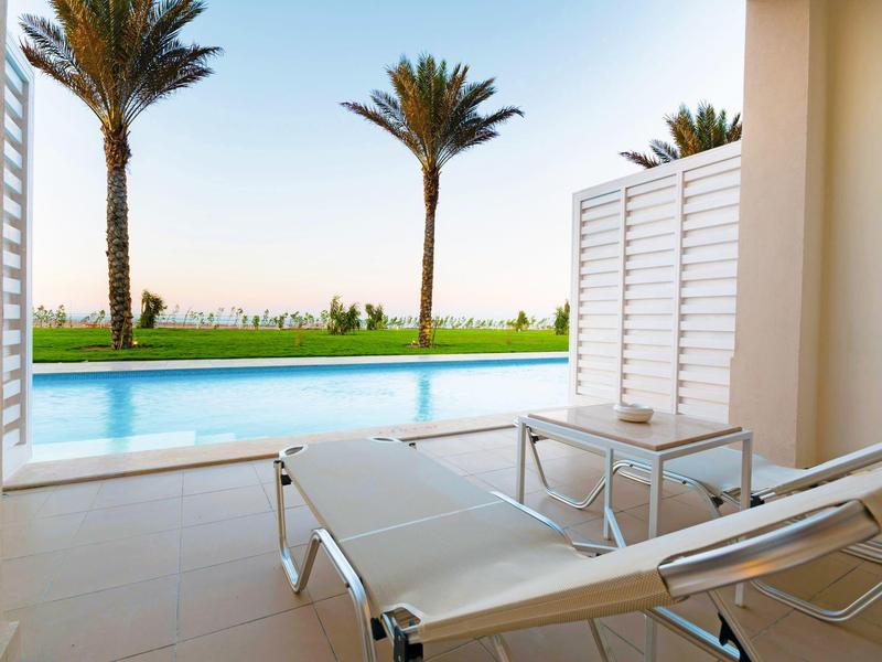 Moderne Terrasse mit Liegestühlen, Pool und zwei Palmen vor grünem Rasen und blauem Himmel.