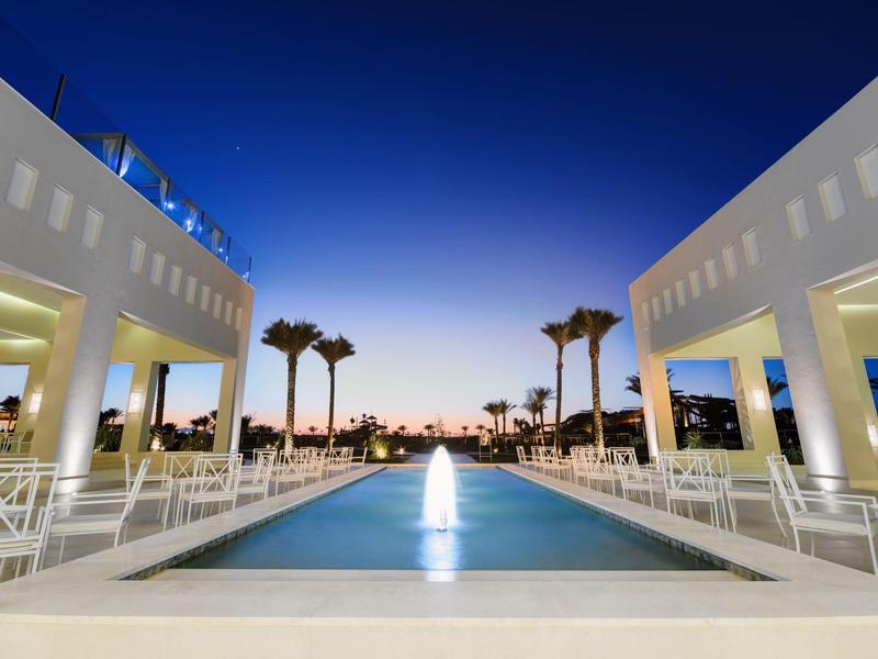 Symmetrischer Pool mit Brunnen zwischen weißen Gebäuden und Palmen bei Sonnenuntergang.