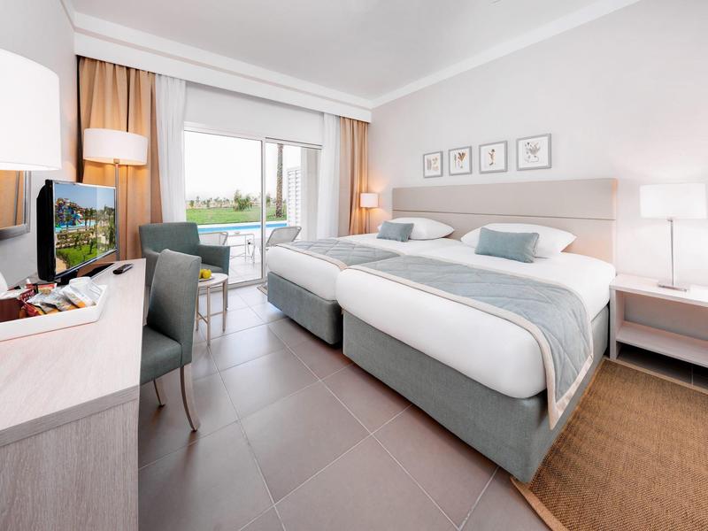 Helles Hotelzimmer mit Doppelbett, Schreibtisch, Stuhl, Fernseher und Balkon mit Grünblick.