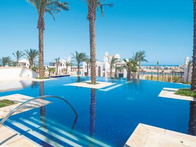 Blauer Pool mit Palmen, weißem Fliesenrand und Blick auf das blaue Meer im Hintergrund.