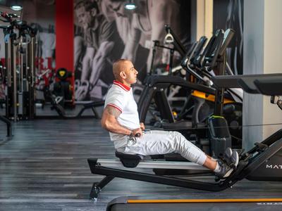 Ein Mann trainiert auf einem Rudergerät in einem modernen Fitnessstudio mit grauem Boden und roten Säulen.