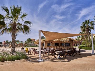 Outdoor-Bereich eines Hotels mit Holzstühlen, Tischen unter einem Sonnensegel und Palmen.