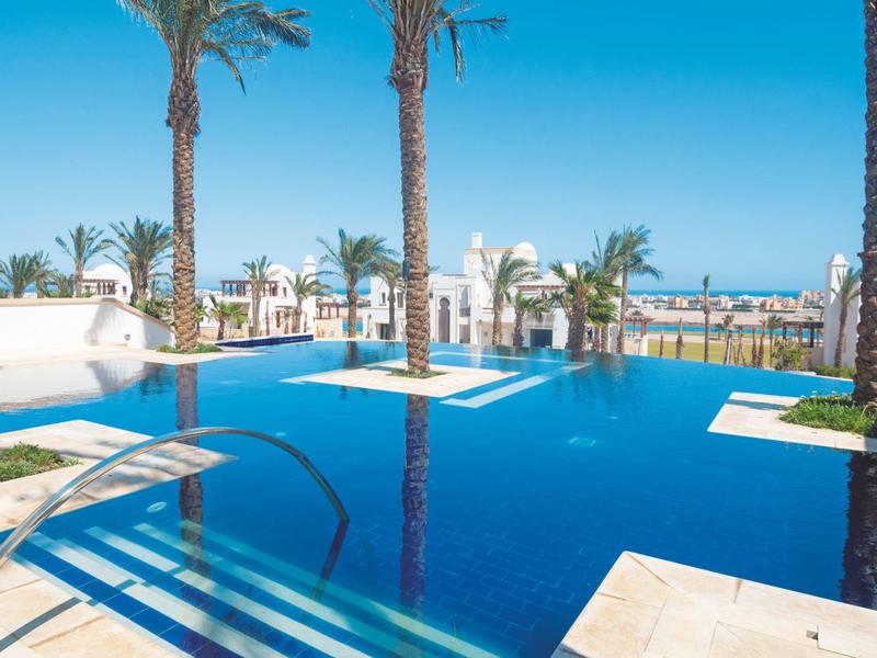Outdoor pool surrounded by palm trees with white buildings and ocean view in the background.