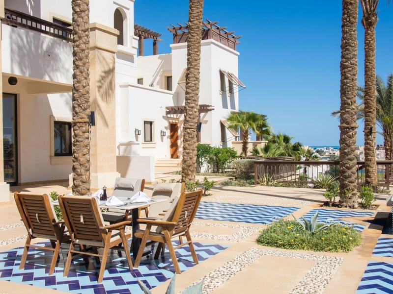 Outdoor terrace with wooden table and chairs, palm trees, and white building under clear blue sky.
