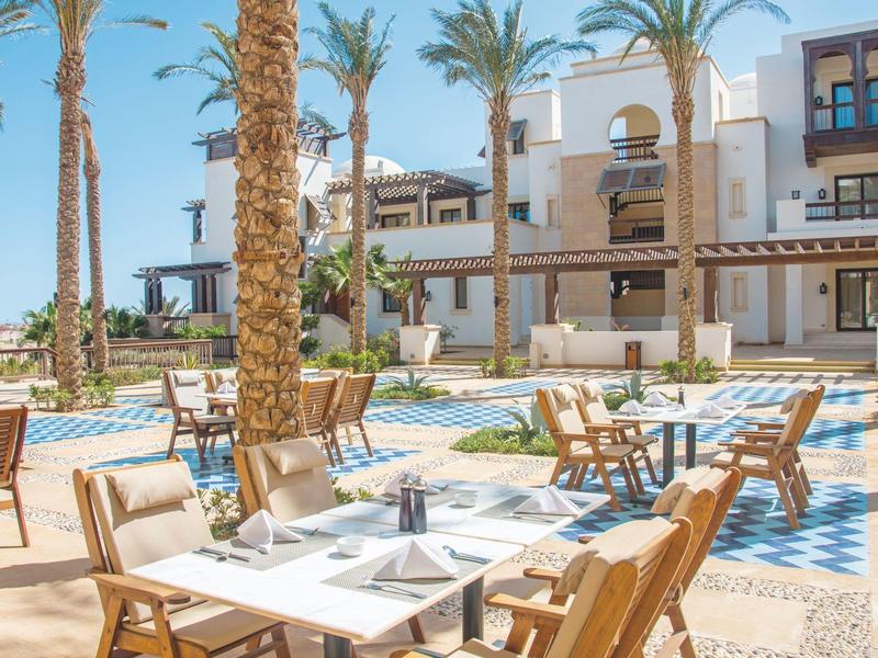 Outdoor dining area with tables and chairs under palm trees near hotel and pool