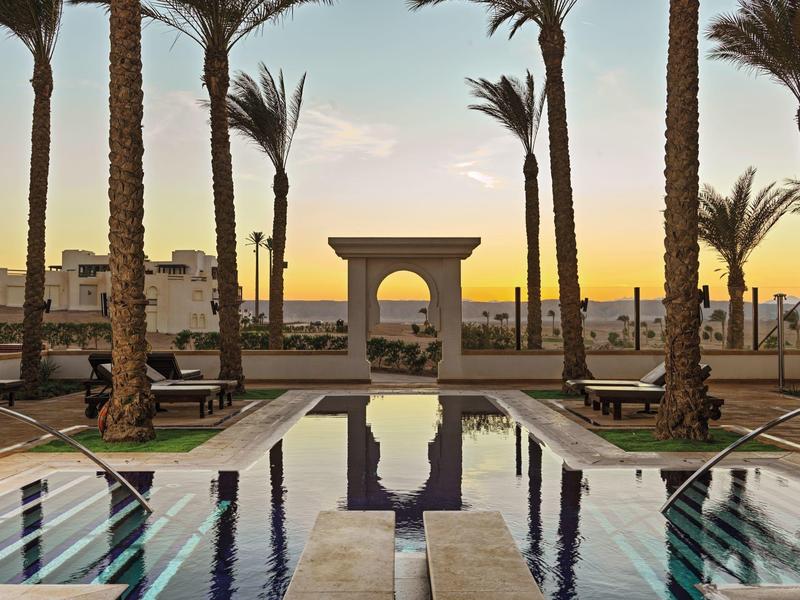 Sunset view of a pool framed by tall palm trees and a stone archway in a tropical resort.