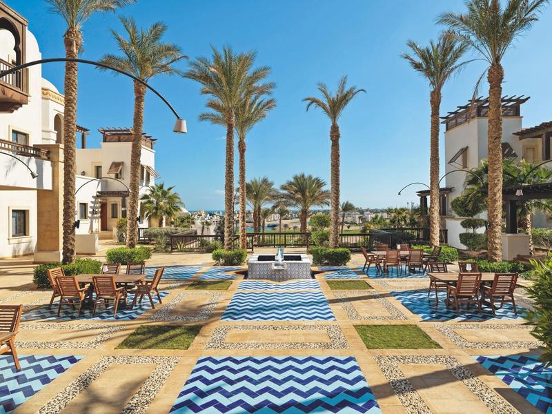 Sunny resort seating area with palm trees and patterned pathways under clear blue sky.