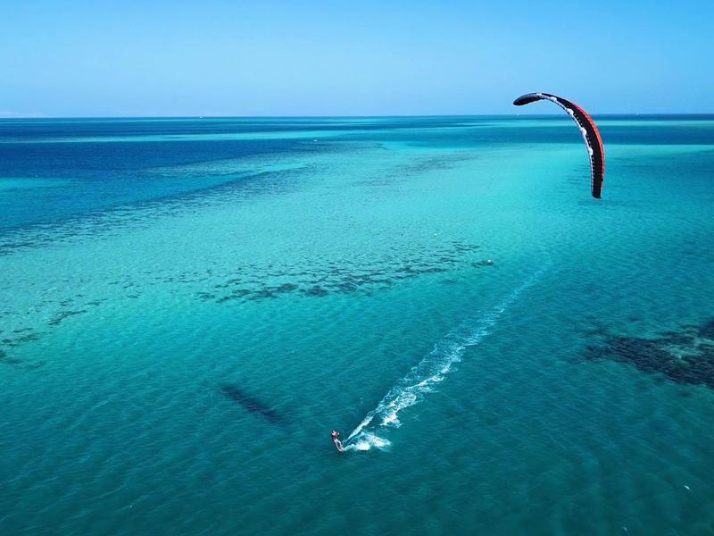 A person kite surfing over clear turquoise ocean waters under a bright blue sky.