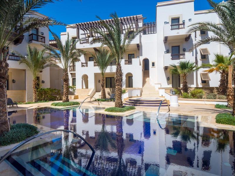 Cozy hotel lounge with plush seating, cushions, plant, and view of outdoor patio with umbrella and bench.