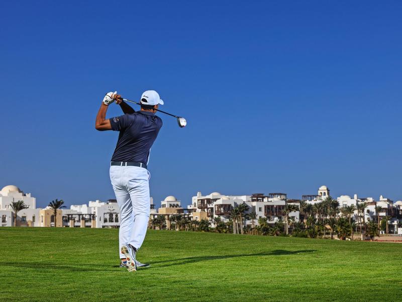Golfer swinging on lush green course with clear blue sky and buildings in distance.