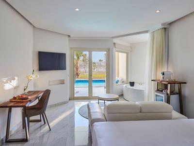 Modernes Hotelzimmer mit Doppelbett, Schreibtisch, Fernseher und Blick auf Pool durch Glaswand.