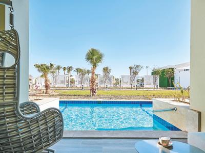 Blick durch Fenster auf Pool mit blauer Wasserfläche, Palmen und Sonne an klarem Himmel.