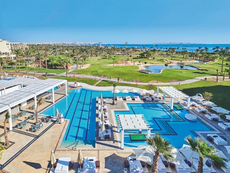 Großer Hotelpool mit Liegestühlen und Blick auf einen Golfplatz und das Meer im Hintergrund.