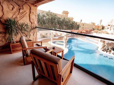 Balkon met uitzicht op zwembad, twee houten stoelen en tafel, rotsmuurdecoratie en blauwe lucht.