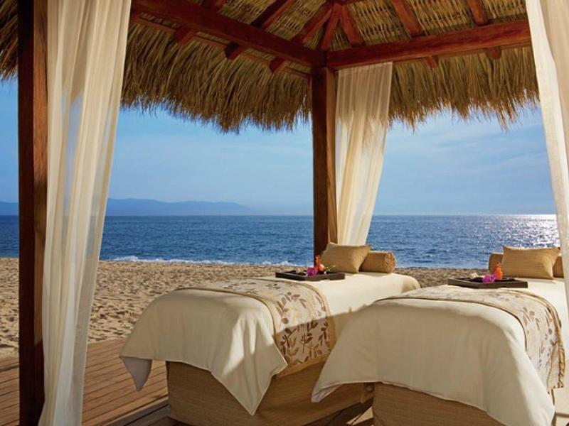 Twee massagetafels onder een rieten dak met uitzicht op het strand en de zee bij zonsondergang.