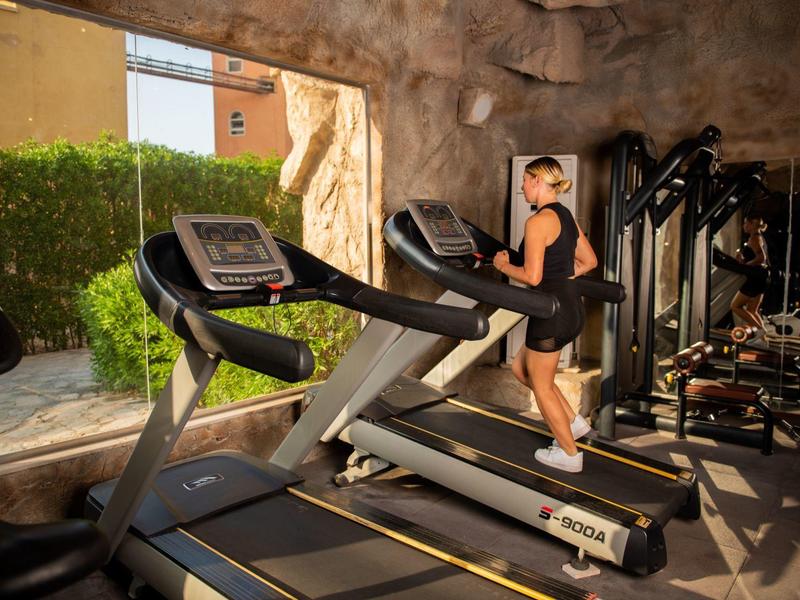 Vrouw die hardloopt op een loopband in een lichte sportschool met ramen en fitnessapparatuur.