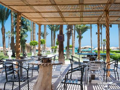 Überdachte Terrasse mit Metalltischen und -stühlen, Palmen und Blick auf blauen Himmel und Meer.