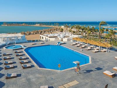 Großer blauer Pool mit weißen Liegen und Palmen, dahinter Meer und bewölkter Himmel.