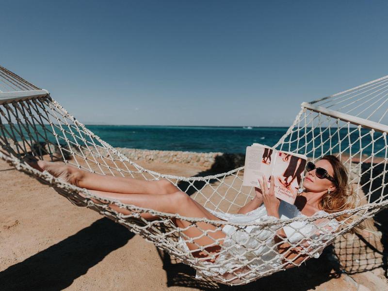 Frau entspannt in Hängematte am Strand mit Buch und Sonnenbrille.