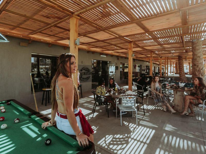 Offener Innenbereich mit Billardtisch, Sitzgelegenheiten und Personen unter einem Holzlatten-Dach.