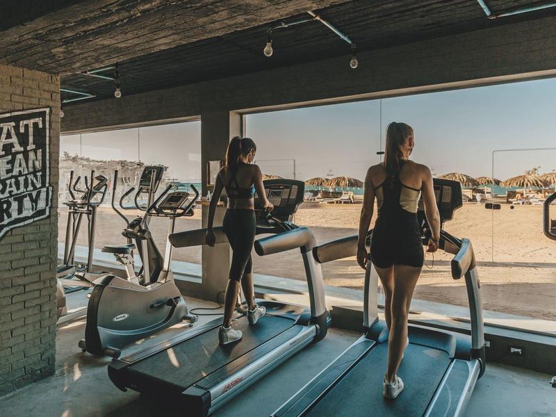 Zwei Frauen laufen auf Laufbändern neben einem Fenster mit Aussicht auf eine sonnige Wüstenlandschaft.