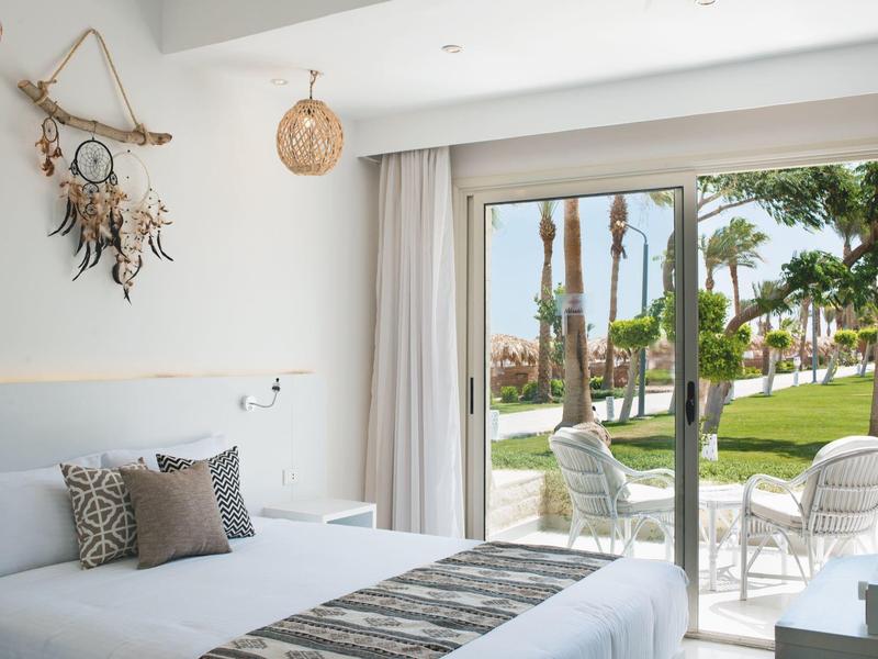 Helles Hotelzimmer mit Doppelbett, Dekoration an der Wand und Blick auf Garten und Terrasse.