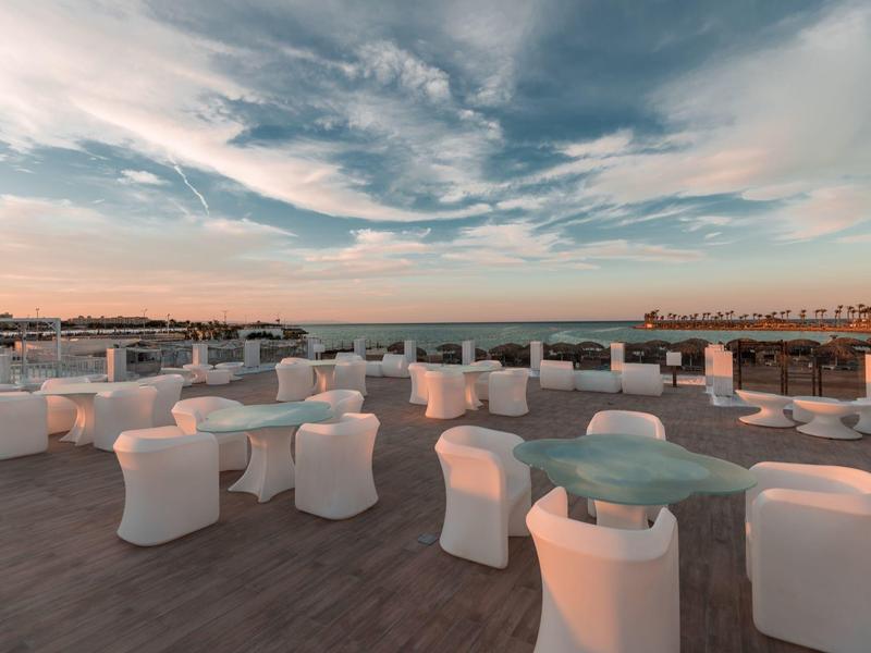 Moderne Dachterrasse mit weißen Möbeln und Glasgeländer unter dramatischem Himmel bei Sonnenuntergang.