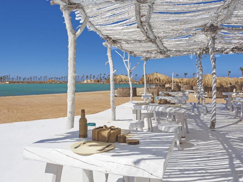 Weißer Strand mit Sonnenschutz und Tischen am ruhigen blauen Meer unter klarem Himmel.