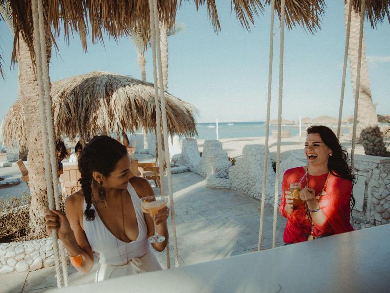 Zwei Frauen sitzen an einer Strandbar mit Strohdach, trinken Cocktails und lachen.