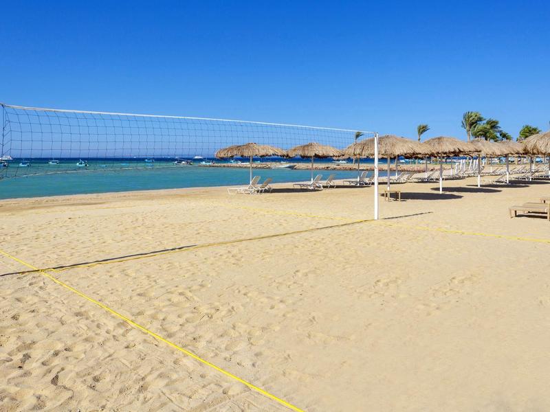 Sandstrand mit Volleyballnetz und Liegestühlen unter Sonnenschirmen am Meer.