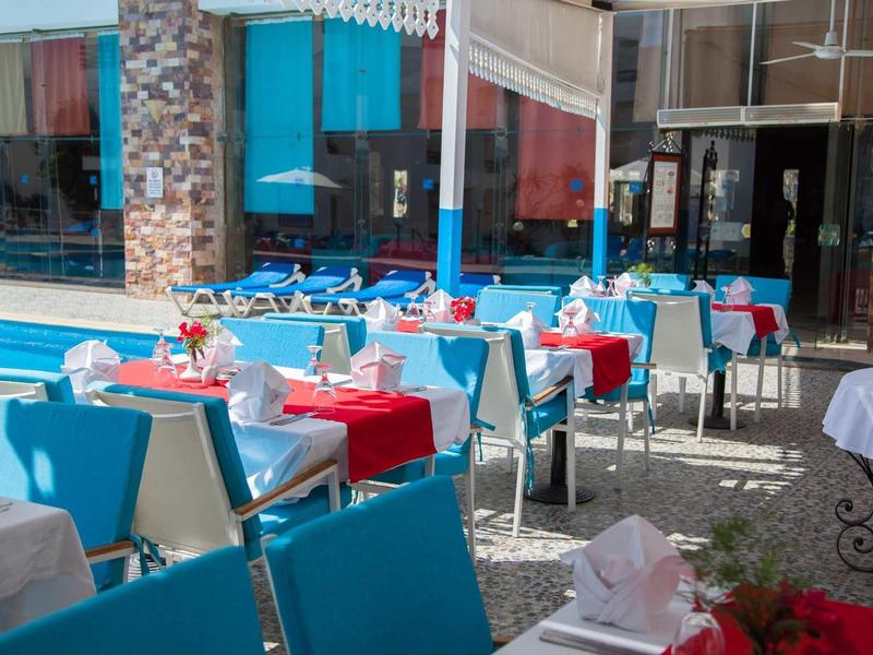 Área de restaurante al aire libre con sillas azul y blanco y manteles rojo y blanco bajo el sol.