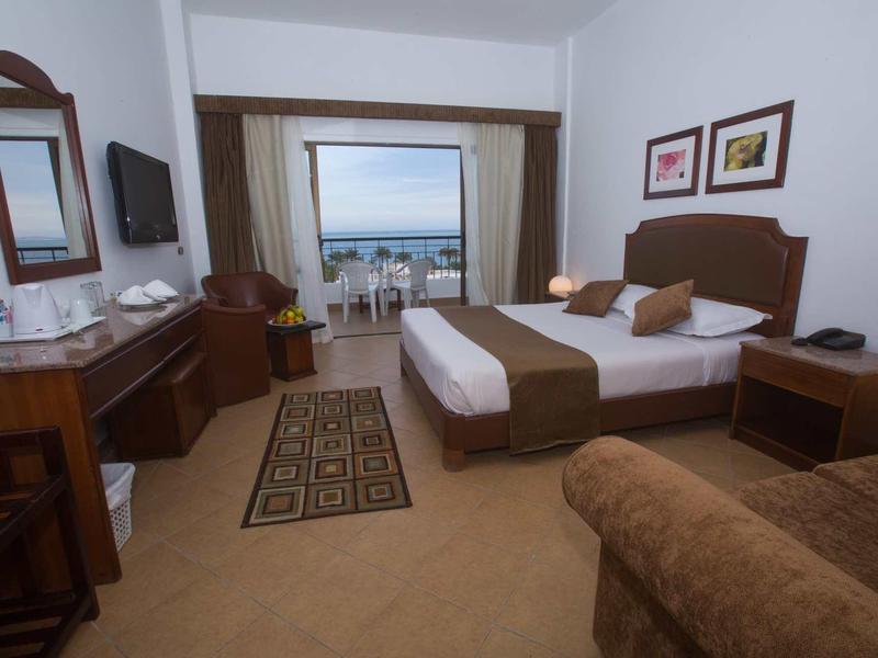 Habitación de hotel luminosa con cama doble, escritorio, televisor por cable y balcón con vista al mar.