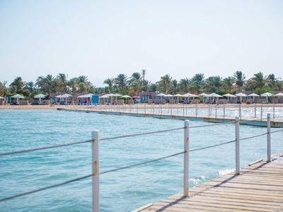 Hölzerner Steg mit Metallgeländer über blauem Meer, Strand mit Palmen und Sonnenschirmen im Hintergrund.