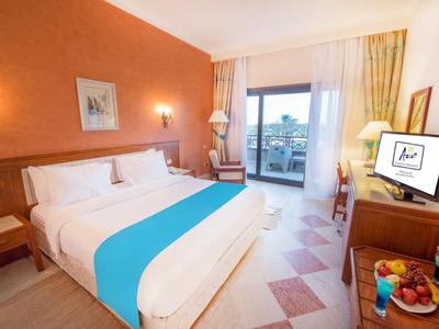 Helles Hotelzimmer mit Doppelbett, türkisfarbener Bettdecke, Balkon und Fernseher.