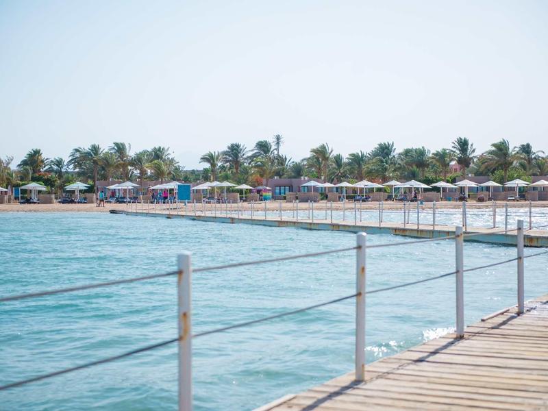 Un ponton en bois au-dessus de l'eau avec une plage et des parasols en arrière-plan.