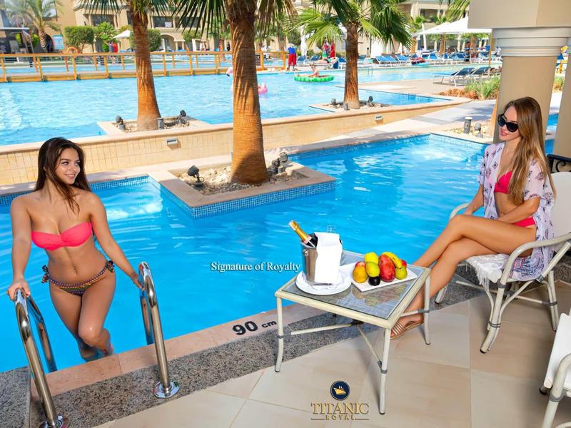 Deux femmes se détendent à la piscine de l'hôtel avec des fruits, des boissons et des palmiers en arrière-plan.